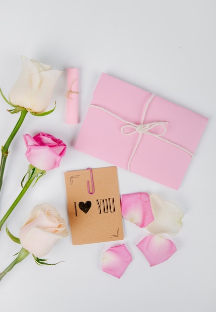 Top view of white and pink color roses with envelope tied with a rope and small postcard with a paper clip on white background