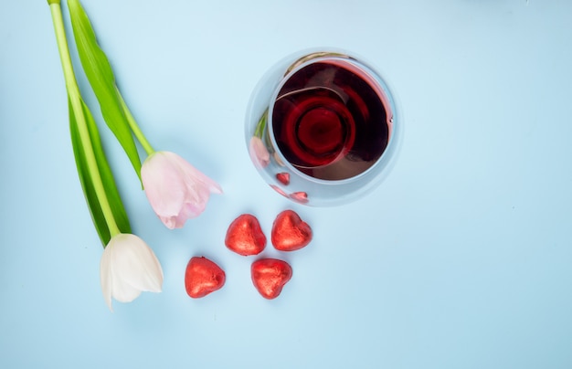 Top view of white and pink color tulip flowers with scattered heart shaped candies in red foil and a glass of wine on blue table