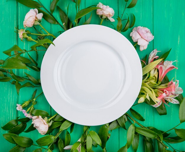 Top view of white plate on light pink flowers with leaf branches on a green surface