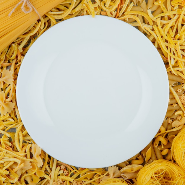 Top view of white plate on raw pasta of different shapes and types as on background