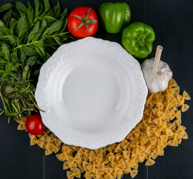 Top view of white plate with raw pasta tomatoes garlic and bell pepper with mint on a black surface