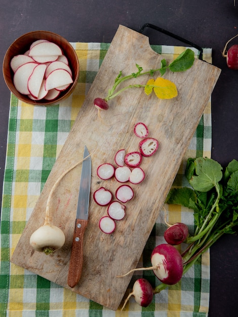 Top view of white and red radishes on cutting board with knife on cloth surface and maroon background