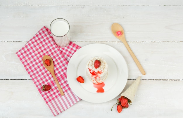 Top view white rice wafers on plate with red gingham tablecloth,strawberries,wooden spoons and milk on white wooden board surface. horizontal