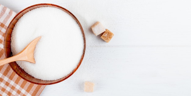 Top view of white sugar in a wooden bowl with a spoon and lump sugar cubes on white background with copy space