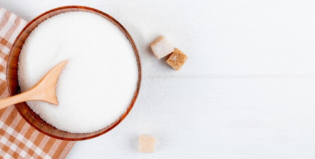 Top view of white sugar in a wooden bowl with a spoon and lump sugar cubes on white background with copy space