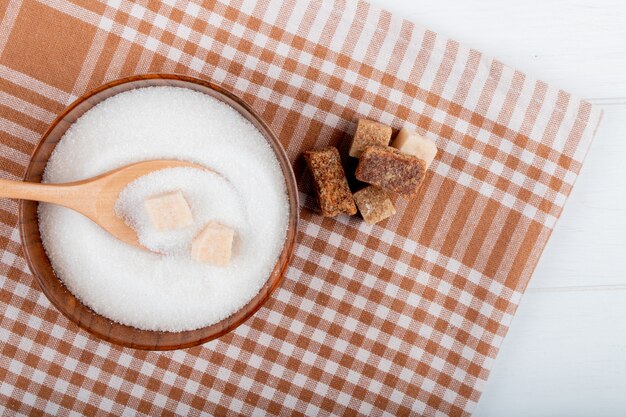 Top view of white sugar in a wooden bowl with a spoon and lump sugar and palm sugar pieces on plaid tablecloth with copy space