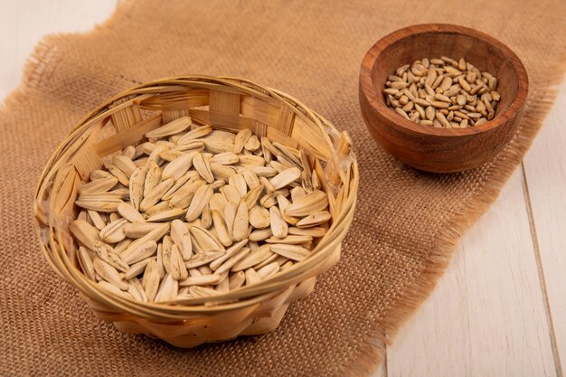 Free photo top view of white sunflower seeds on a bucket on a sack cloth with shelled sunflower seeds on a wooden bowl on a beige wooden table