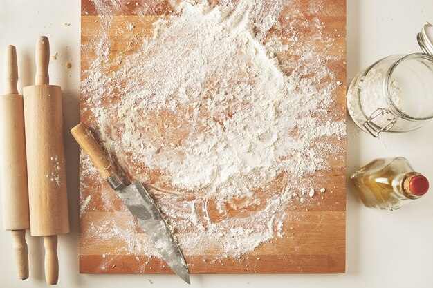 Top view on white table with isolated wooden board with knife, two rolling pins, bottle olive oil, transparent jar with flour. Presentation cooking process