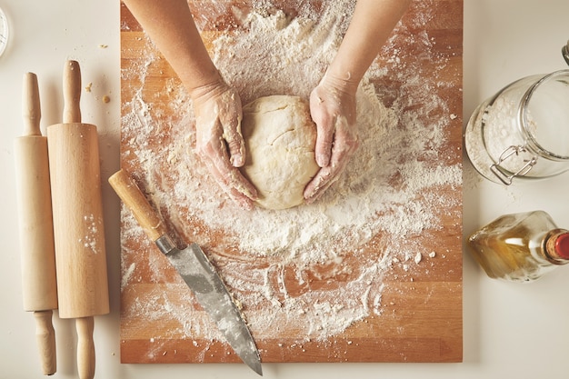 Top view on white table with isolated wooden board with knife, two rolling pins, bottle olive oil, transparent jar with flour Woman hands hold prepared dough for pasta or dumplings