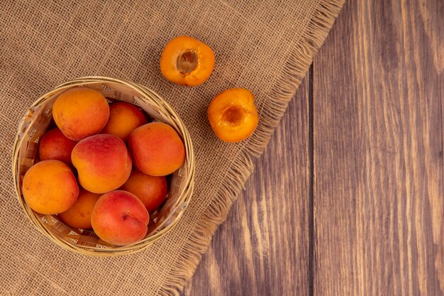 Top view of whole apricots in basket and half cut one on sackcloth on wooden background with copy space