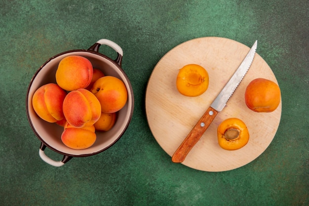 Top view of whole apricots in bowl and half cut one with knife on cutting board on green background