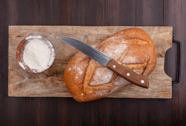 Top view of whole crusty bread and bowl of flour with knife on cutting board on wooden background