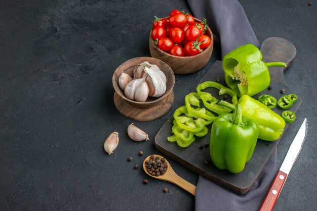 Top view of whole cut chopped green peppers on black wooden cutting board knife on towel tomatoes garlics in bowls on black distressed surface