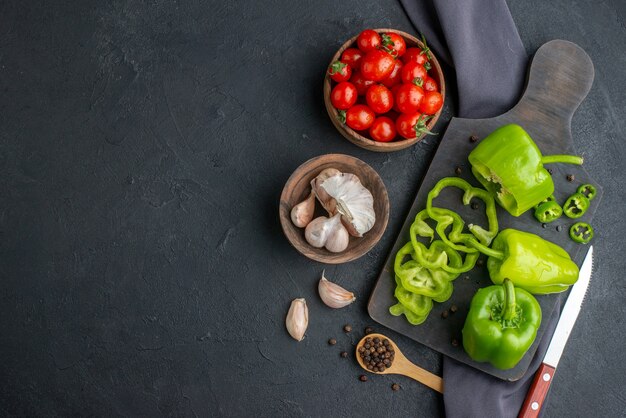 Top view of whole cut chopped green peppers on wooden cutting board tomatoes in bowl garlics on dark color towel on the left side on black surface