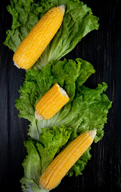 Top view of whole and cut cooked corns with lettuce on black surface