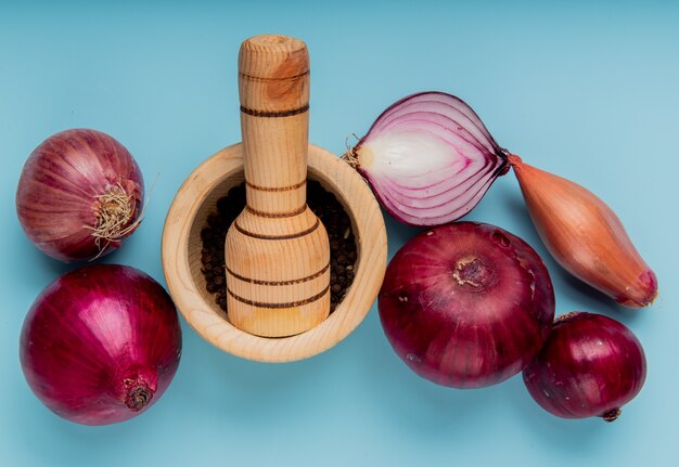 Top view of whole and cut onions and black pepper seeds in garlic crusher on blue surface