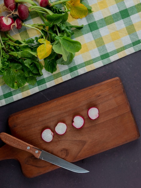 Top view of whole and cut radishes on cloth and cutting board with knife on maroon background