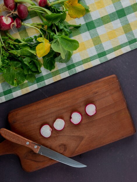 Top view of whole and cut radishes on cloth and cutting board with knife on maroon background
