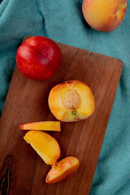 Top view of whole cut sliced peach with knife on cutting board and whole peach on blue cloth surface