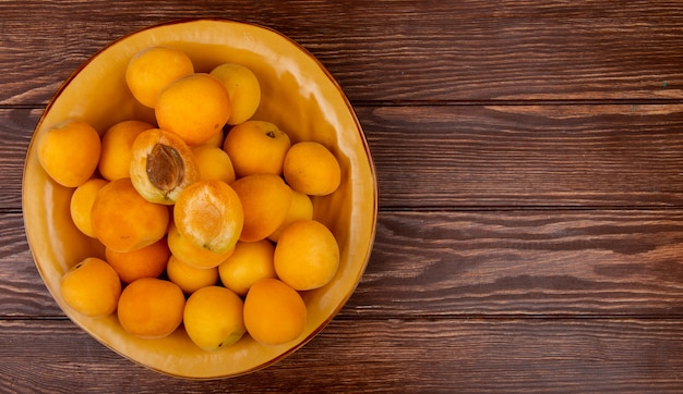 Top view of whole and half cut apricots in bowl on wooden background with copy space