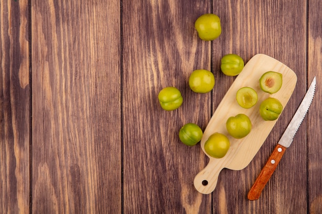 Top view of whole and half cut plums on cutting board with knife on wooden background with copy space