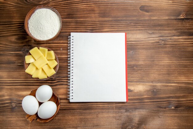 Top view whole raw eggs with flour notepad and cheese on brown table egg dough pastry flour dust