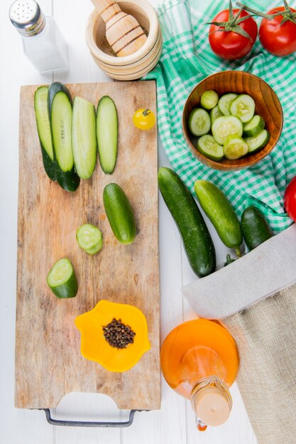 Top view of whole sliced and cut cucumbers and black pepper on cutting board with tomatoes bowl of cucumber slices and cucumbers on cloth with salt garlic crusher melted oil on wooden surface