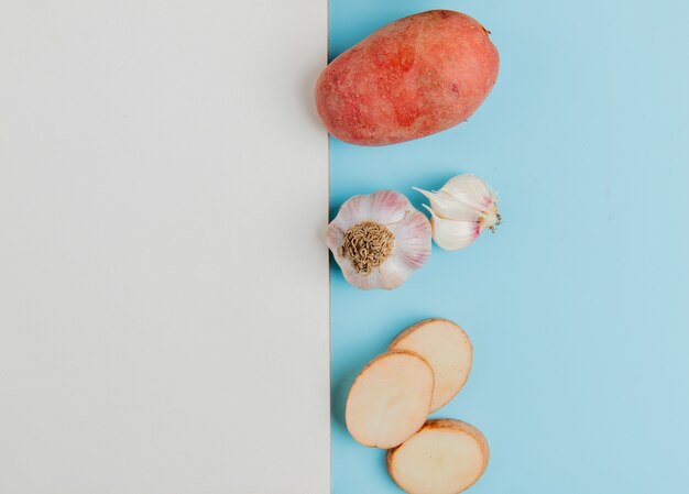 Top view of whole and sliced potatoes with garlic and note pad on blue surface with copy space