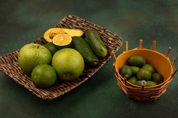 Top view of a wicker tray of healthy foods such as green apples limes avocado and cucumber with feijoas on a bucket on a green surface