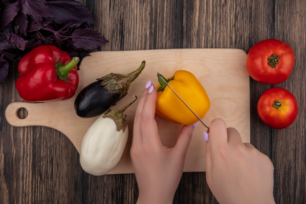 Top view  woman cuts colored bell pepper with eggplants on a cutting board with tomatoes and basil on a wooden background