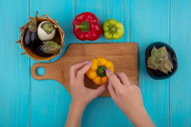 Top view  woman cuts colored bell peppers on a cutting board with a knife with white and black eggplants in a basket on a turquoise background