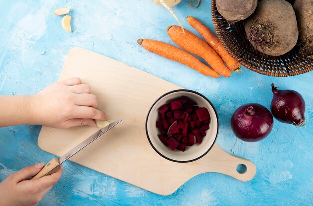 Top view woman cuts garlic on a board with chopped beets in a cup with carrots and red onions on a table