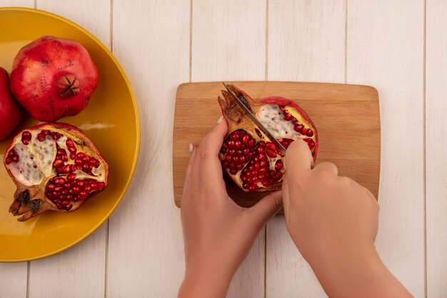 Top view woman cuts half a pomegranate on a cutting board with a whole on a plate on a white wall