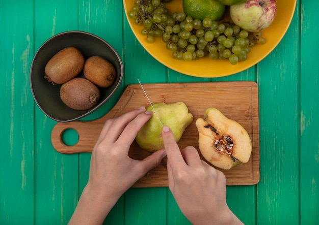 Foto gratuita vista dall'alto la donna taglia la pera su un tagliere con kiwi e mandarini su una parete verde