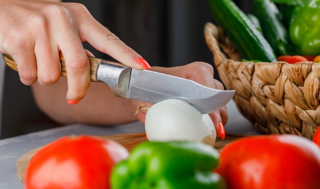 Top view woman cutting onion into half on cutting board with knife, green pepper, cucumber, salt on gray surface