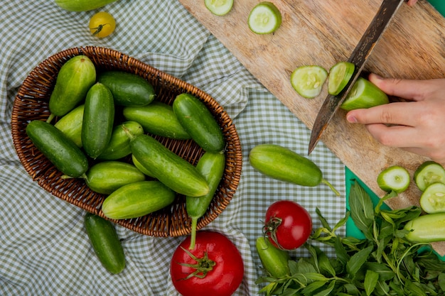 Top view of woman hand cutting cucumber with knife on cutting board with other ones in basket tomatoes and mint on plaid cloth surface