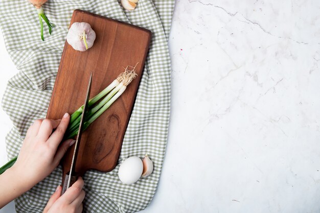 Free photo top view of woman hand cutting green onion on cutting board with garlic and egg on white background with copy space