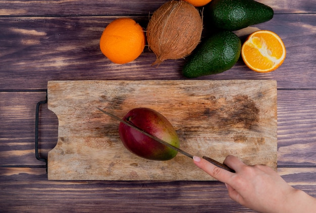Взгляд сверху манго ручной резки женщины с ножом на разделочной доске и авокадо мандарина кокоса на деревянной предпосылке