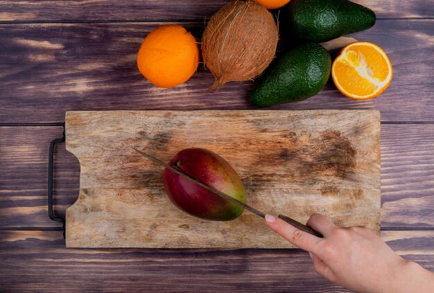 Top view of woman hand cutting mango with knife on cutting board and coconut tangerine avocado on wooden background