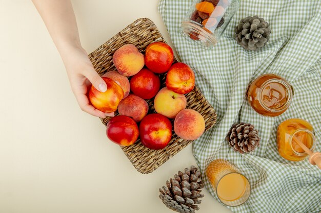 Top view of woman hand holding basket plate of peaches with plum jam peach juice raisins and pinecones on cloth on white surface