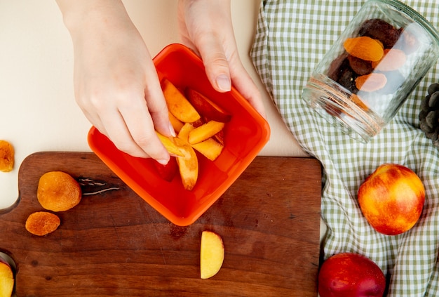 Top view of woman hand holding bowl of peach slices with dried plums on cutting board and peaches with lying jar of raisins on cloth and white surface