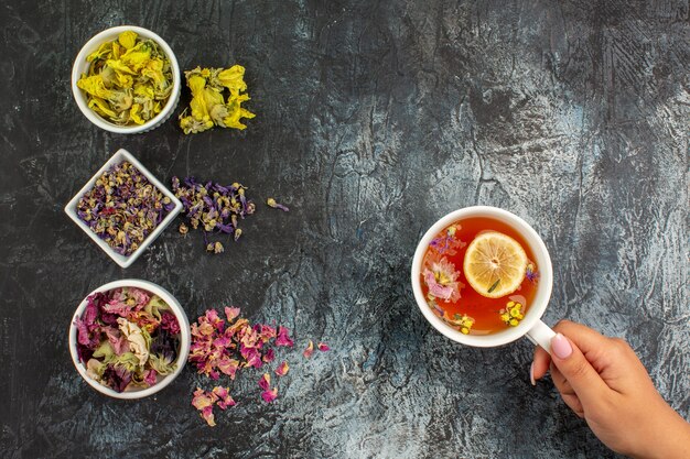 Top view of woman hand holding a cup of herbal tea and three bowls of dry flowers on grey ground