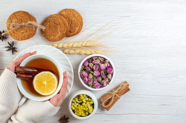 Top view of woman hand holding a cup with black tea with cinnamon lime lemon and various herbals cookies on the right side on white background