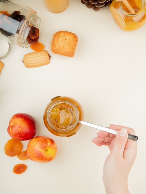 Top view of woman hand holding glass jar of peach jam with peaches raisins cupcake on white surface