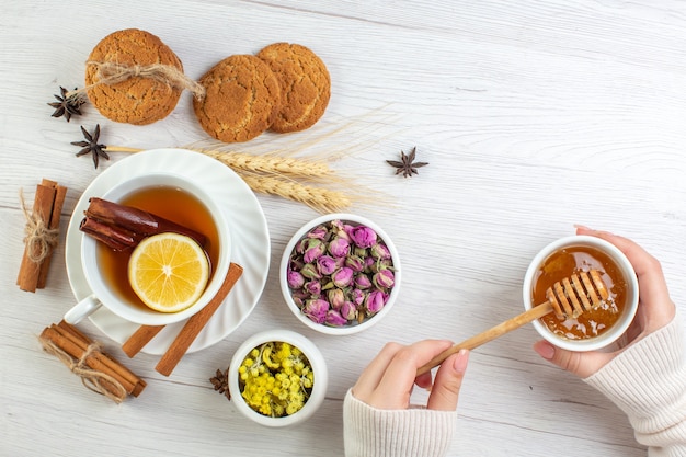 Top view of woman hand holding honey in a pot with black tea with cinnamon lime lemon and various herbals cookies on white background