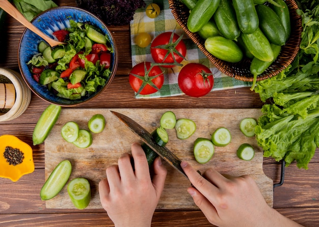 Top view of woman hands cutting cucumber with knife on cutting board with vegetable salad lettuce tomato black pepper on wooden surface