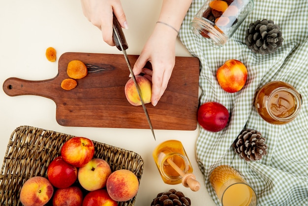 Top view of woman hands cutting peach with knife and dried plums on cutting board with jams and juice raisins and pinecones around on white surface