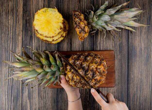 Top view of woman hands cutting pineapple with knife on cutting board with sliced pineapple on wooden table