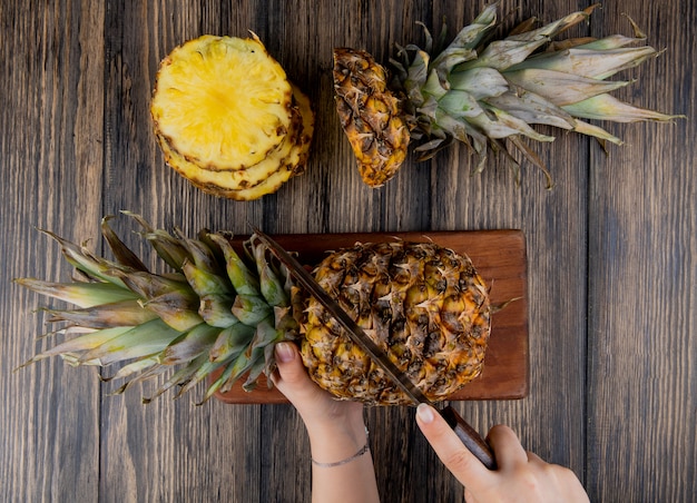 Top view of woman hands cutting pineapple with knife on cutting board with sliced pineapple on wooden table