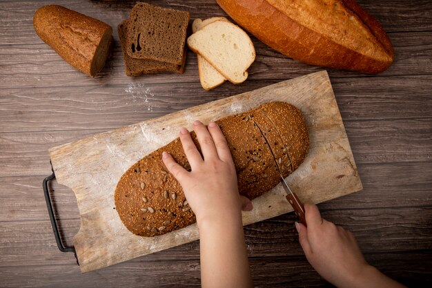 Top view of woman hands cutting sandwich bread on cutting board and other breads on wooden background
